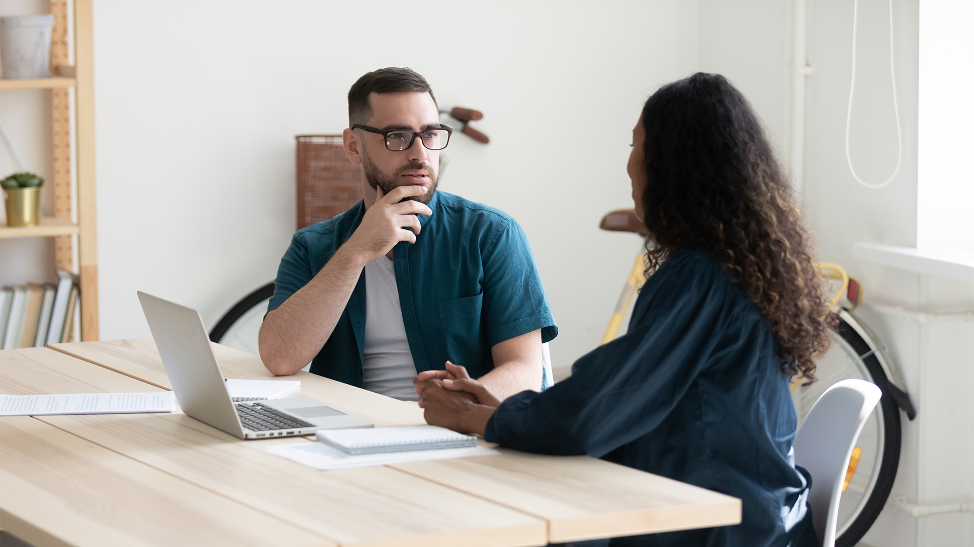Two office workers sitting at a desk talking and an open Windows 11 Pro PC open between them, with a bookcase and notebooks on the desk
