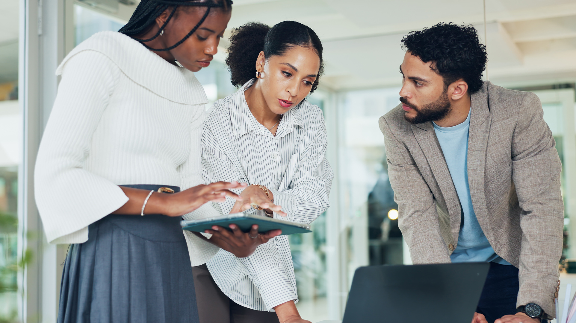 Three office workers standing together over a desk, one worker holding a 2-in-1 Copilot+ PC tablet, all discussing onscreen content in an office setting