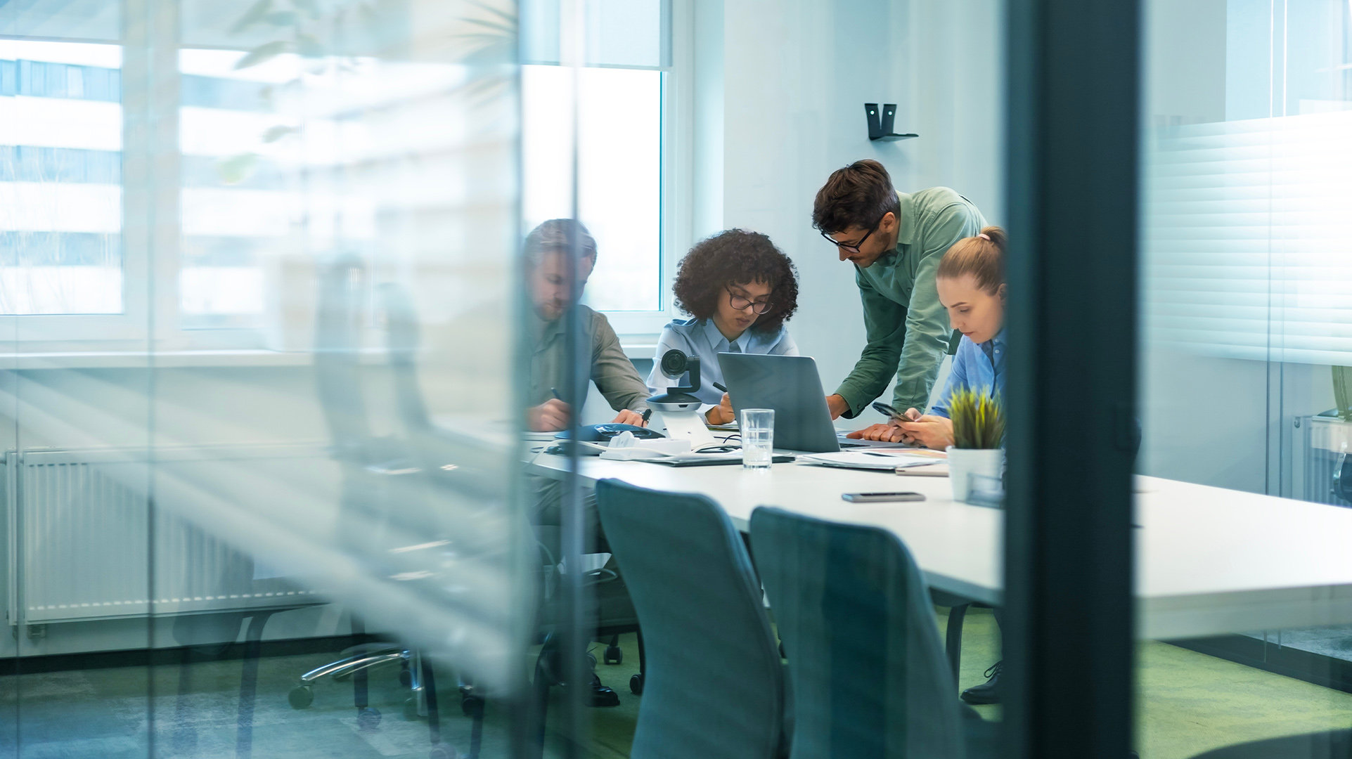 Four office workers meeting in an enclosed large meeting room with glass walls discussing onscreen information from an open Windows Pro PC, with a plant on the table, and other papers