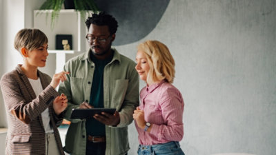Three office workers standing together in a meeting looking on as one worker holds and open Windows 11 Pro device and a bookcase in the background