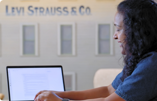 An office worker sitting in a large, bright meeting space presenting data on an open Windows 11 Copilot+ PC with the Levi Strauss and Company logo in the background