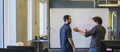 Dve osobe stoje ispred bele table i razgovaraju o dijagramima i beleškama u opremljenoj kancelariji.
