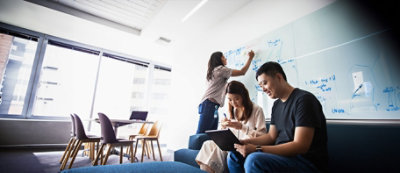 Tri osobe rade u dobro osvetljenoj kancelariji; jedna piše na beloj tabli, a druge dve sede na kauču