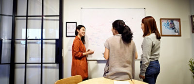 Tri žene u opremljenoj kancelariji stoje oko bele table i razgovaraju o idejama. Jedna žena piše na beloj tabli 