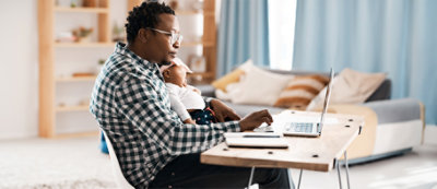 A man holding a baby while working on a laptop.