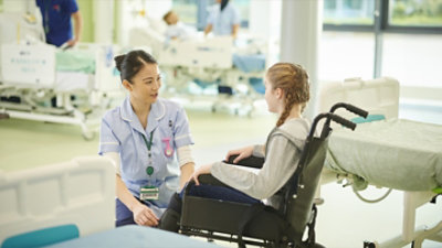 Medicinska sestra u uniformi kleči pored mlade devojke u invalidskim kolicima i razgovara sa njom na bolničkom odeljenju sa krevetima u pozadini.
