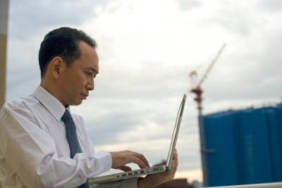 Person in business attire working on a laptop outdoors, with industrial structures and a crane in the background.
