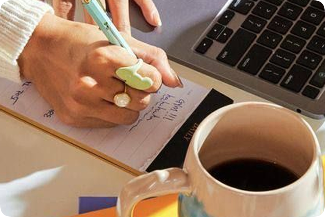 Image of a person writing on a notepad next to their laptop and a cup of coffee