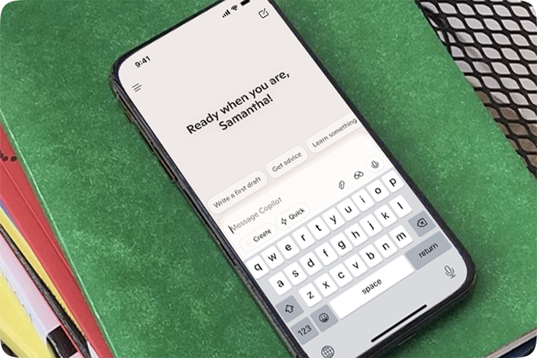 Image of a smartphone on colorful file folders showing the Copilot mobile interface and keyboard with “Ready when you are, Samantha”