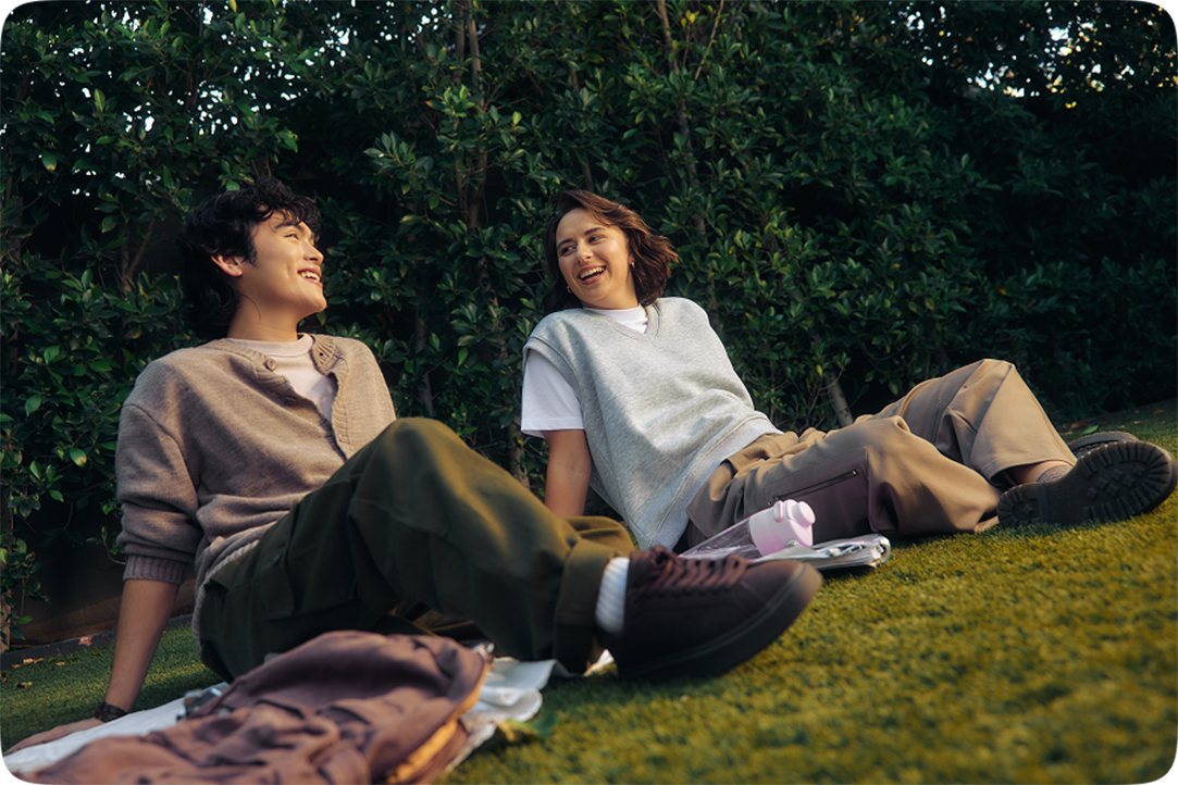 Two people reclining on grass with notebooks and backpacks beside them in a leafy outdoor setting