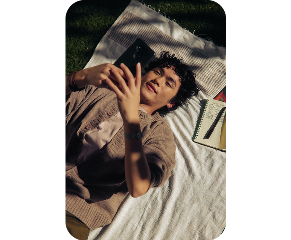 Person lying on a blanket outdoors, holding a phone beside an open notebook and pen