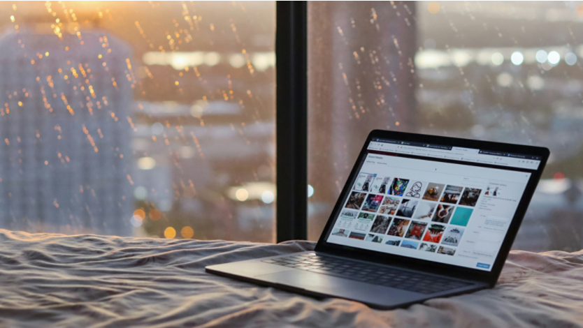 A Surface laptop on a bed against a large window during a rainy sunset