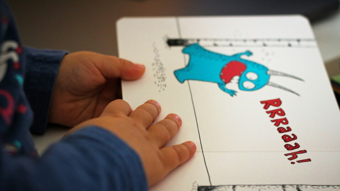 A child’s hand rests on the page of a picture book with an image of a blue monster on a white background