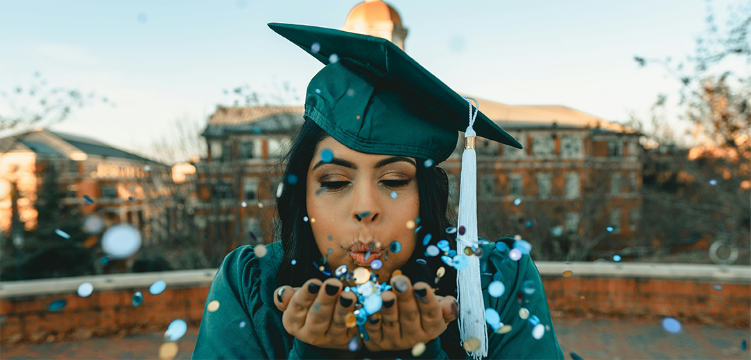A college graduate wearing a green cap and gown and blowing confetti into the camera