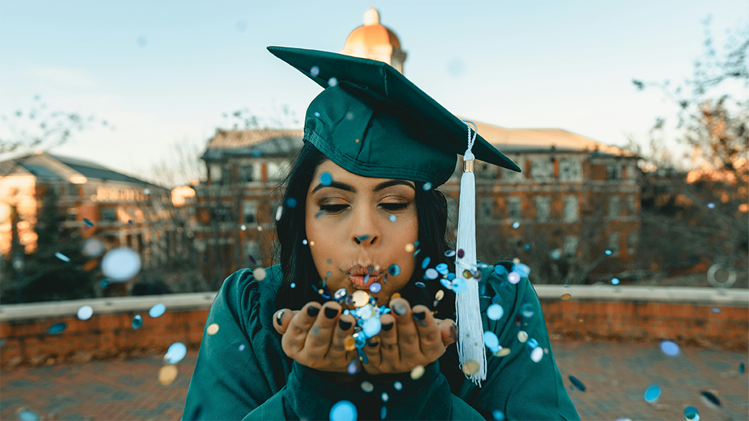 A college graduate wearing a green cap and gown and blowing confetti into the camera
