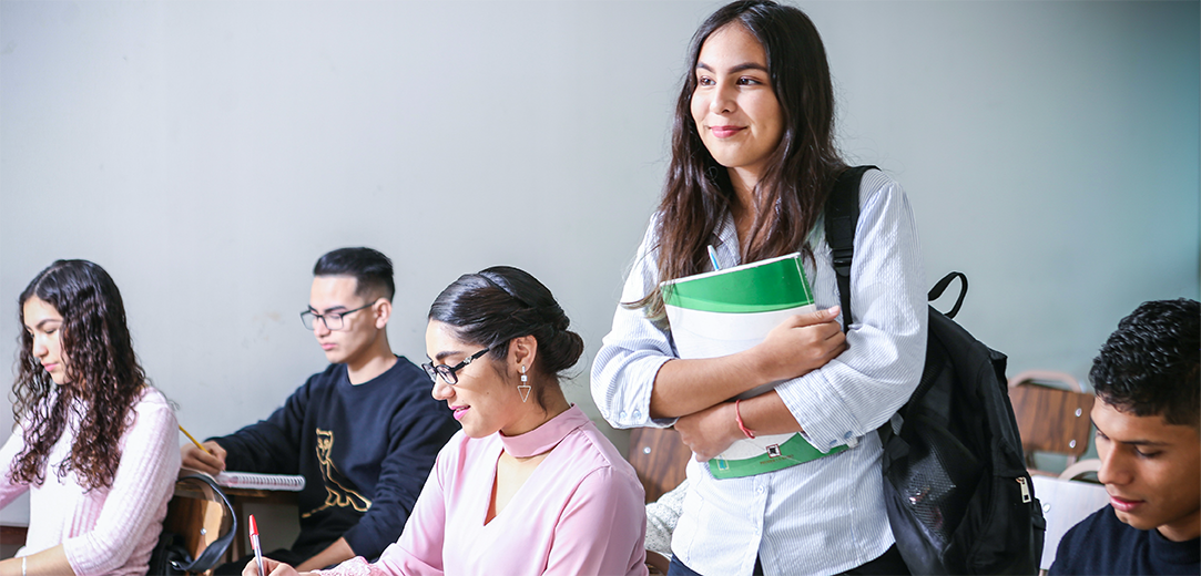 A college student carrying a green and white textbook and wearing a black backpack finds her seat in a classroom filled with other students writing in notebooks
