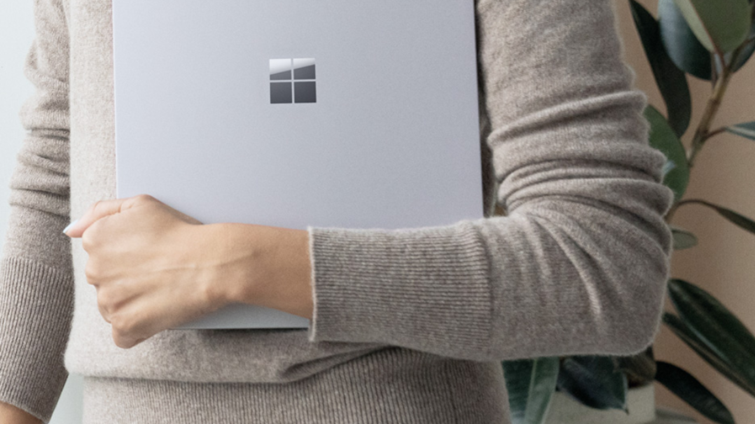 A cropped image of a woman holding a closed Surface device with one arm