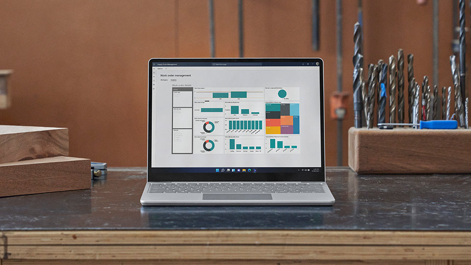 A front-facing Surface Laptop Go 2 is observed on a desk in a wood shop setting. Drill bits are placed nearby