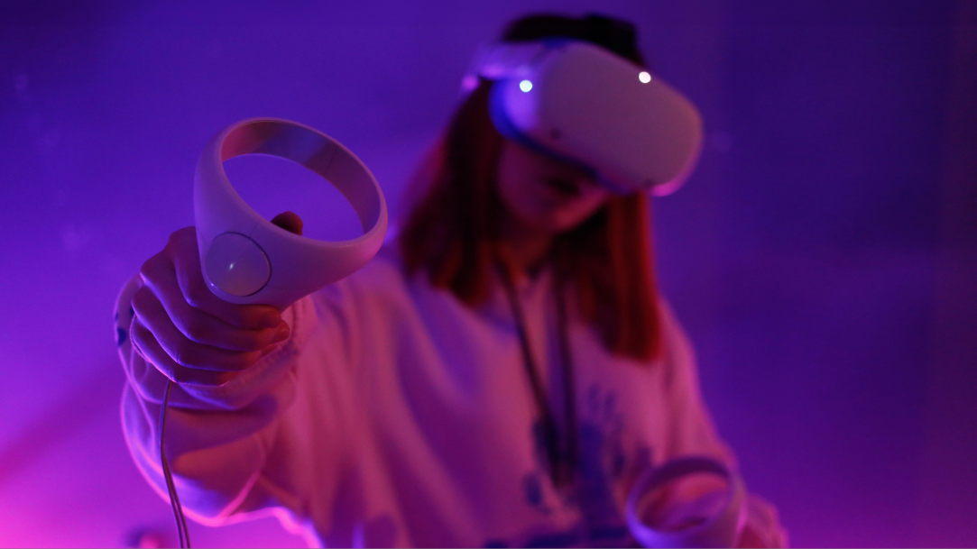 A girl with a white virtual reality headset holds matching controllers in purple and pink gradient lighting