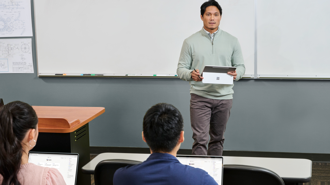 A male standing at the front of a classroom with a whiteboard behind him, holding a Surface Pro 12-inch