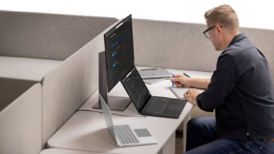 A man sitting at a desk with a black Windows laptop, platinum Surface laptops, and a monitor