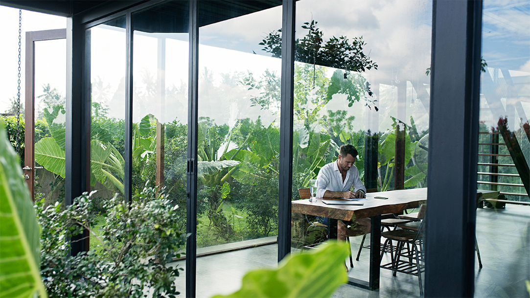 A man sitting at a table and writing in front of lush garden