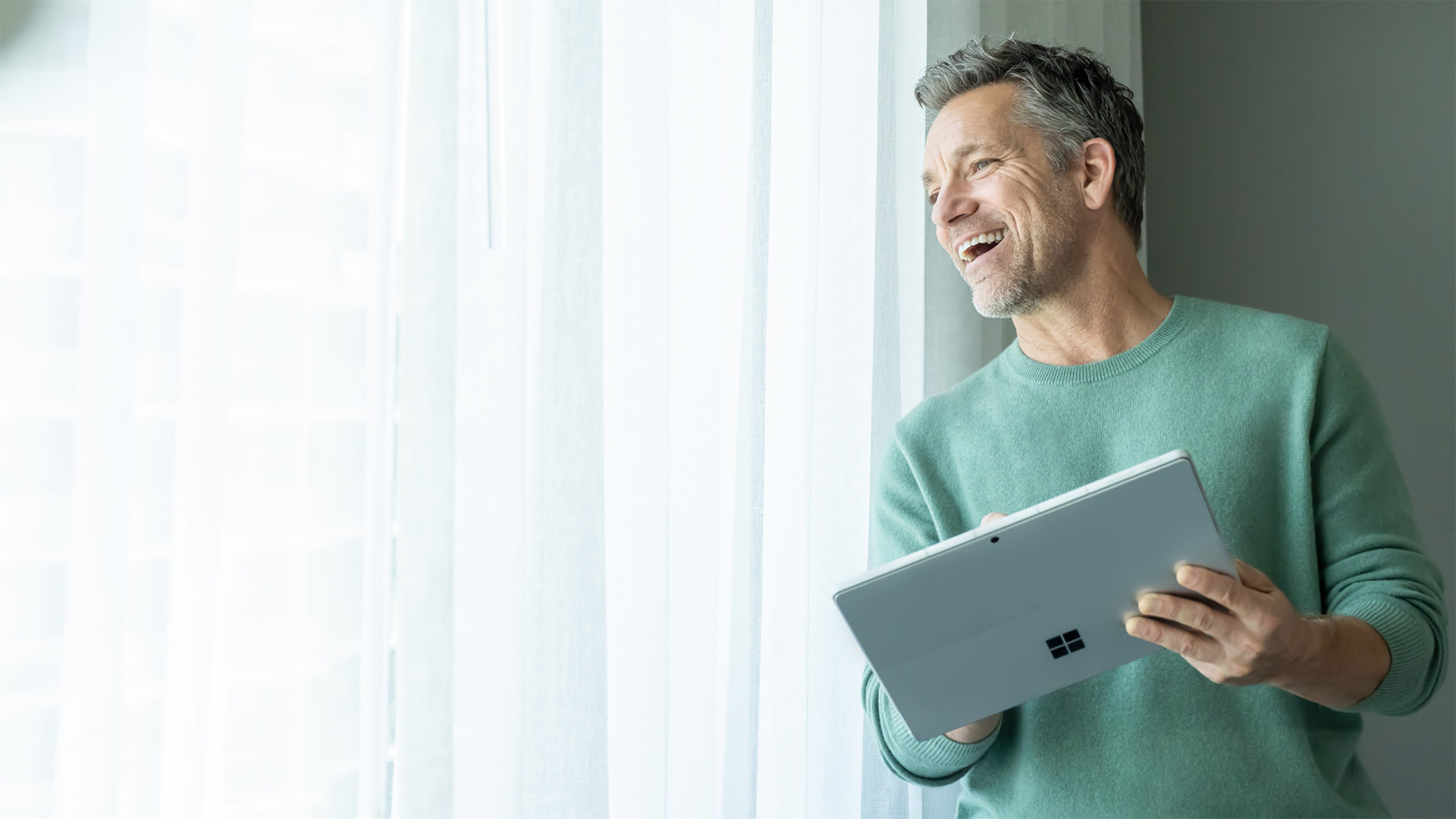 A man standing, smiling and looking out a window while holding a Surface device