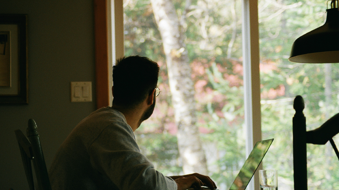 A man using a laptop from his home office and looking out of a window