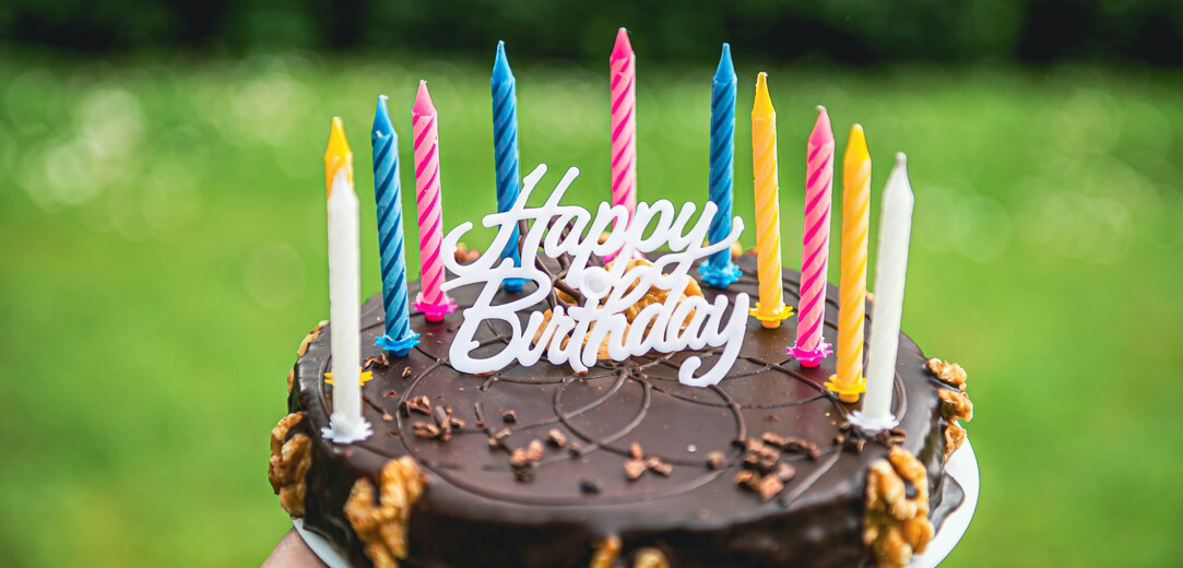 A person holding a round chocolate cake that says “Happy Birthday” with candles around the border