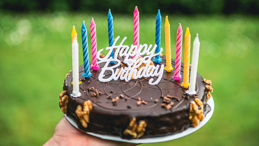 A person holding a round chocolate cake that says “Happy Birthday” with candles around the border