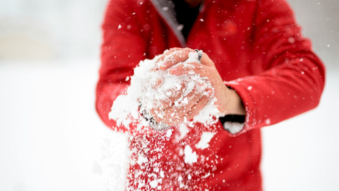 A person in a red jacket forming a snowball in their bare hands