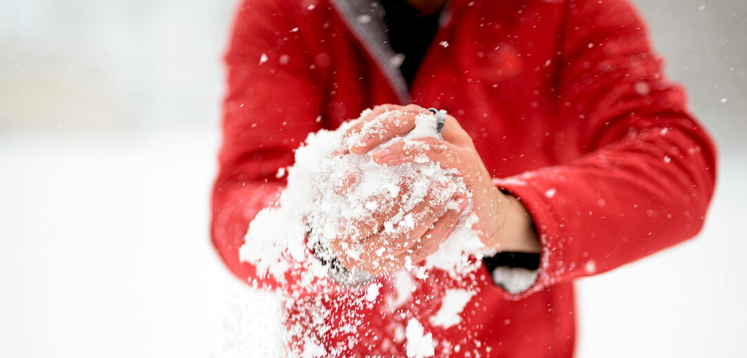 A person in a red jacket forming a snowball in their bare hands