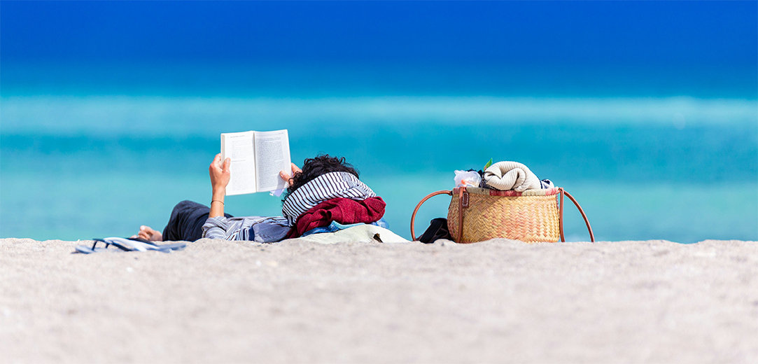 A person reads a book on the beach while lying down on a towel with a woven beach bag next to them