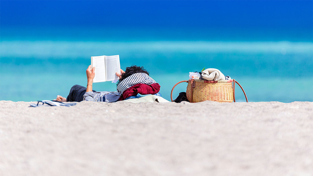 A person reads a book on the beach while lying down on a towel with a woven beach bag next to them