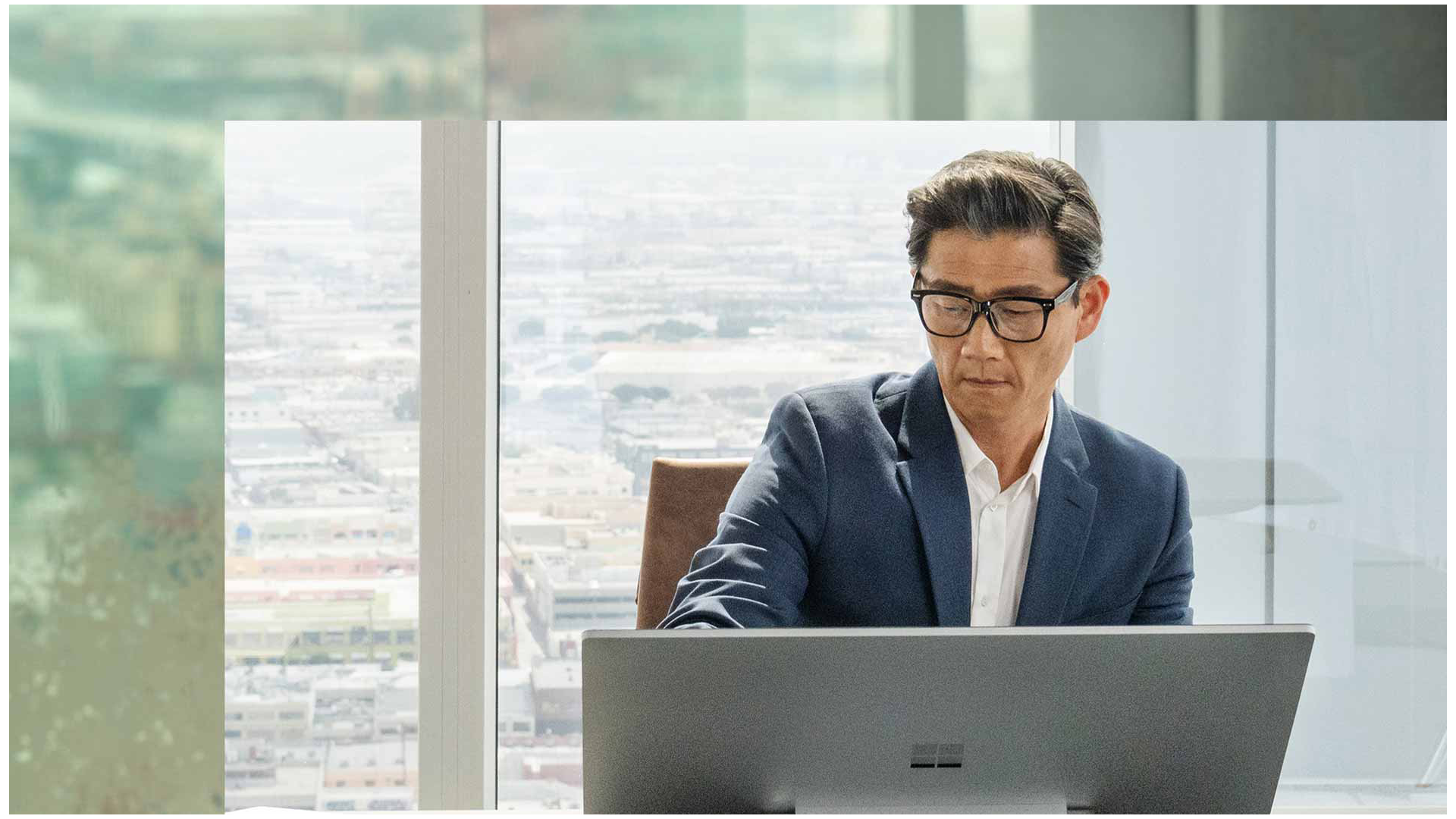 شخص يجلس أمام جهاز Surface Studio 2+‎ في مبنى مكاتب شاهق. تتراءى المدينة في الخلفية.
