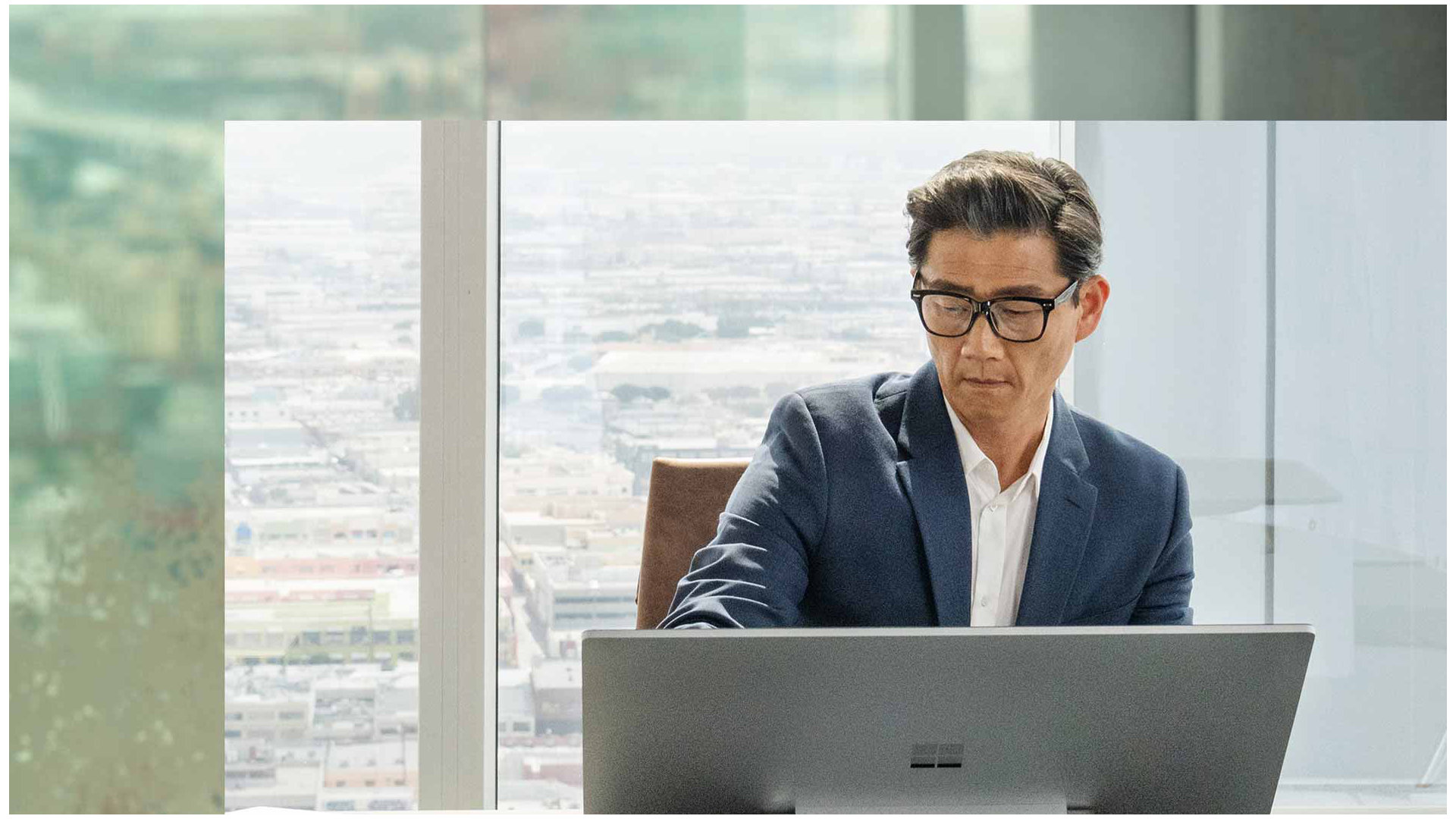 En person sitter ved sin Surface Studio 2+ i en høy kontorbygning. Byen kan ses i bakgrunnen.
