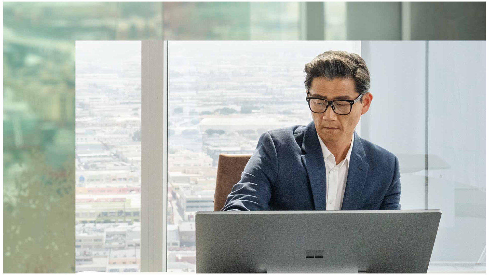 Una persona sentada detrás de su dispositivo Surface Studio 2+ en un edificio de oficina. Se observa una ciudad en el fondo.