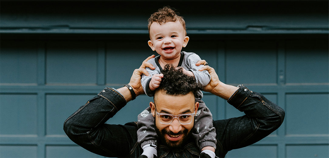 A smiling dad with his smiling baby on his shoulders in front of a dark teal background