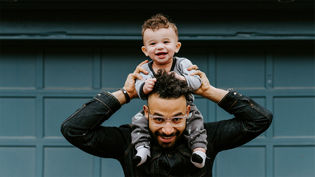 A smiling dad with his smiling baby on his shoulders in front of a dark teal background