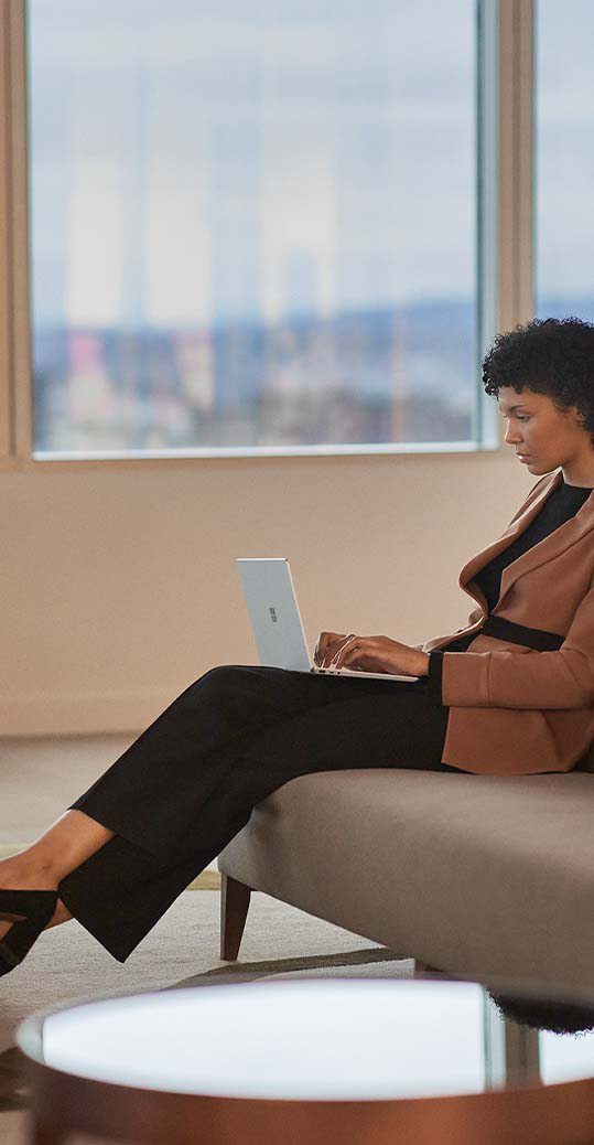 A woman is observed sitting on a sofa in an office setting while typing on her Surface Laptop Go 2