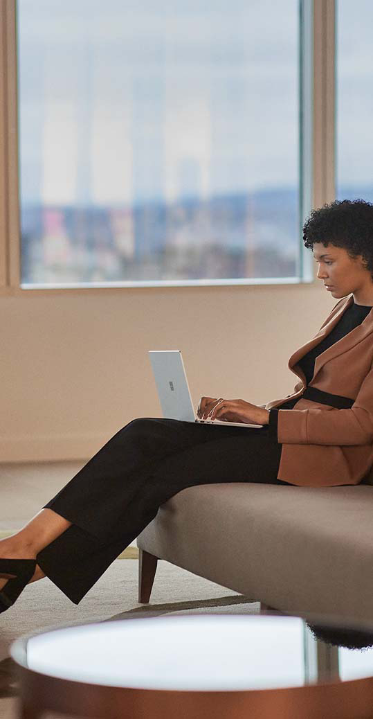 En kvinde ses siddende på en sofa i et kontormiljø og skriver på sin Surface Laptop Go 2