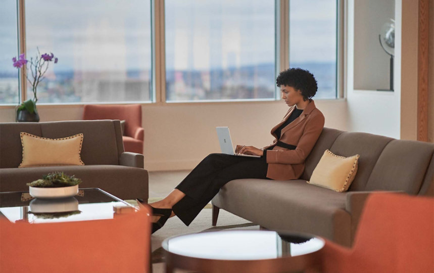 A woman is observed sitting on a sofa in an office setting while typing on her Surface Laptop Go 2