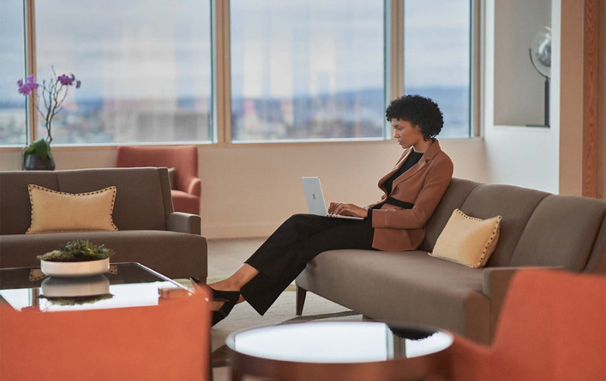 Se observa una mujer sentada en un sofá en una oficina, y que escribe en su Surface Laptop Go 2