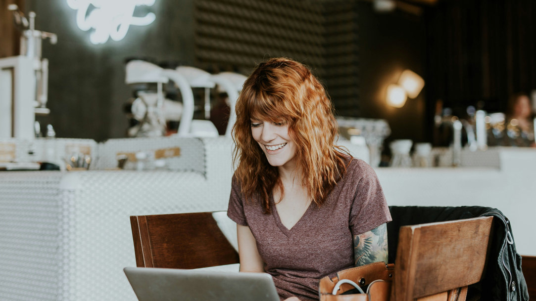 A woman sitting in a cafe with her laptop open smiling while she does her work or video chats with a friend