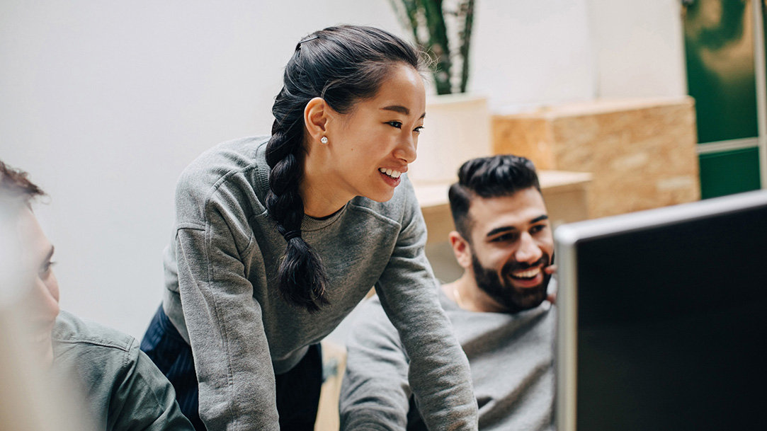 A woman standing and two men sitting as they smile and look intently at a monitor, all wearing gray sweatshirts