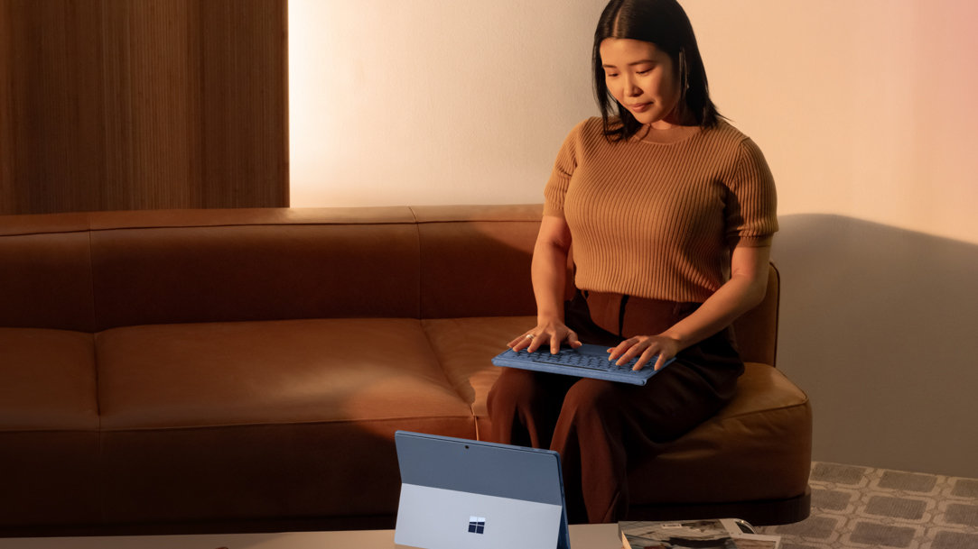 A woman sitting on a sofa while using the detached Surface Pro Flex Keyboard keyboard with a Surface Pro 11th Edition