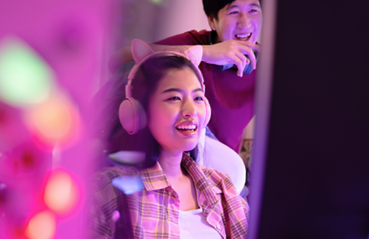 A woman wearing pink headphones gaming on her computer with her friend cheering for her