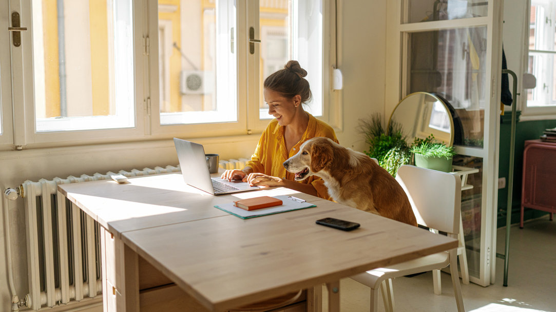 A woman working on a computer on a table with her dog sitting next to her