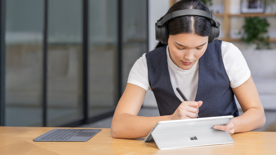 A young woman using a Surface Pro in tablet mode and writing with a Surface Slim Pen, with a Surface Pro keyboard detached and off to the side of the table