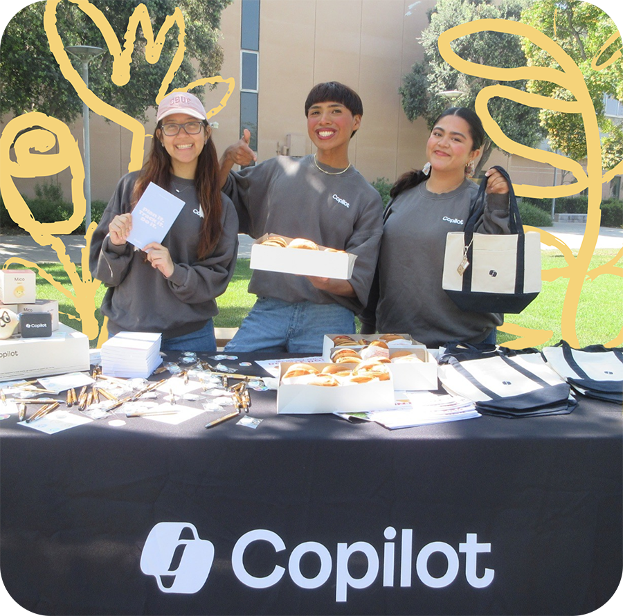 Three Copilot Student Advocates smiling at a Copilot brand table, holding up Copilot merch and a box of donuts.